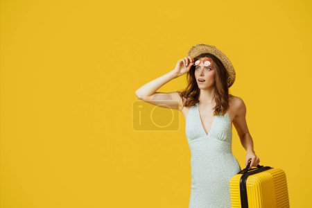 Téléchargez les photos : Young lady in sunglasses and summer hat holding suitcase and looking at copy space on yellow studio background. Woman getting ready for vacation - en image libre de droit