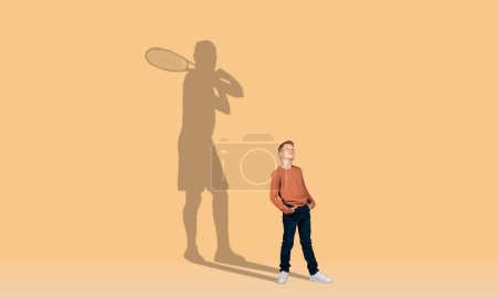 Téléchargez les photos : Mignon petit garçon rousse avec les mains dans les poches et l'ombre d'un joueur de tennis adulte avec raquette derrière lui sur fond de mur coloré studio, enfant rêvant de sport professionnel, espace de copie, collage - en image libre de droit