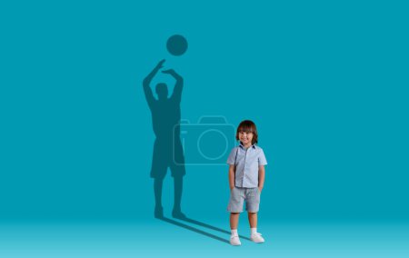 Téléchargez les photos : Cute cheerful smiling little boy with hands in pockets and shadow of adult basketball player behind him on blue studio wall background, kid dreaming about professional sports, copy space, collage - en image libre de droit