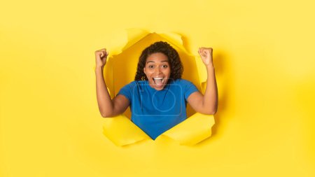 Foto de Mujer negra llena de alegría animando con los puños cerrados, celebrando la victoria, mirando a la cámara con emoción, posando en un agujero roto de fondo amarillo, panorama - Imagen libre de derechos