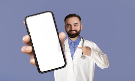 Photo for Smiling Male Doctor Pointing At Big Blank Smartphone In His Hand, Handsome Physician Man In Uniform Demonstrating Empty Cellphone With White Screen At Camera, Recommending Medical App, Mockup - Royalty Free Image