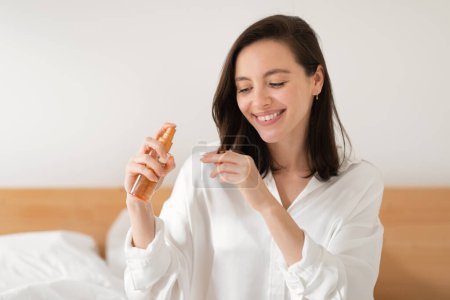 Téléchargez les photos : Smiling young european lady sits on white bed, applies spray on hair, enjoys daily procedures in bedroom interior, copy space. Good morning, wake up and beauty care, treatment and lifestyle at home - en image libre de droit