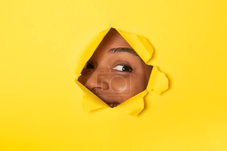Photo for Young african american lady peeking out of a hole in yellow paper background, look aside through torn hole with one eye, free copy space - Royalty Free Image