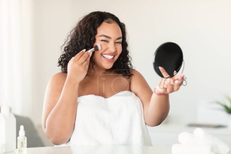 Téléchargez les photos : Daily makeup concept. Excited plus size lady applying blush with makeup brush and smiling at camera, holding mirror, sitting in bedroom. Beautiful woman using new cosmetics - en image libre de droit
