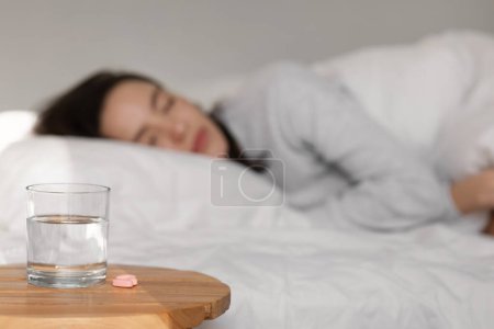 Téléchargez les photos : Triste jeune femme européenne fatiguée ment, dormir sur un lit blanc, se concentrer sélectivement sur un verre d'eau et des pilules sur la table à l'intérieur de la chambre. Problèmes de sommeil, stress, dépression, insomnie et vitamines à la maison - en image libre de droit