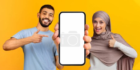 Téléchargez les photos : Couple islamique joyeux pointant vers Big Blank Smartphone et montrant pouce vers le haut, Heureux Arabe Homme Et Femme Publicité Nouvelle application mobile ou site Web, Posant sur fond jaune, Maquette, Collage - en image libre de droit