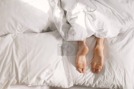 Photo for Legs of young european female lies on white comfortable bed under blanket, wakes up, enjoy free time at night, top view, sun flare, cropped. Lazy good morning, rest and relaxation at home, health care - Royalty Free Image