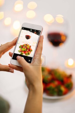 Foto de Manos femeninas tomando fotos de copa de vino tinto y plato lleno de fresa en la mesa festiva decorada con velas encendidas, mujer compartiendo fotos en las redes sociales mientras cena romántica, utilizando el teléfono celular - Imagen libre de derechos