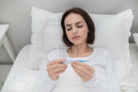 Foto de Upset young sick brunette woman with fever checking her temperature with electronic thermometer, lying in bed at home, lady got sick, copy space. Cold, flu, coronavirus concept - Imagen libre de derechos