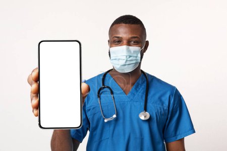 Photo for Black Doctor Wearing Medical Mask Holding Big Smartphone With Blank Screen, Young African American Male Physician Demonstrating Cellphone With Copy Space For Mobile Advertisement, Collage, Mockup - Royalty Free Image