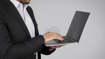 Téléchargez les photos : Homme d'affaires méconnaissable en costume tenant l'ordinateur portable dans les mains tout en se tenant isolé sur fond gris, tir recadré de l'entrepreneur masculin tapant sur le clavier, l'envoi de courriels aux partenaires - en image libre de droit