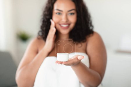 Téléchargez les photos : Heureuse femme en surpoids noire tenant un pot de crème de peau bio, appliquant un hydratant sur le visage après la douche, debout enveloppé dans une serviette, mise au point sélective - en image libre de droit