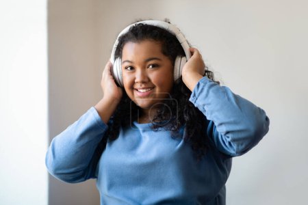 Photo for Portrait of happy young plus size curly woman in homewear listening to her favourite songs music while having fun at home, using brand new wireless headphones, smiling at camera, copy space - Royalty Free Image