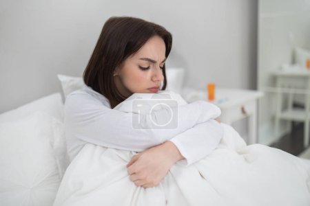Téléchargez les photos : Jeune femme brune malheureuse malade assise au lit à la maison, dame a attrapé froid, grippe ou coronavirus, souffrant de crampes menstruelles ou de maux de tête, médicament sur la table de chevet, espace de copie - en image libre de droit