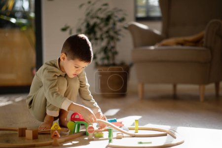 Foto de Busy caucasian little kid builds city, plays road and cars, enjoy free time in kindergarten, preschool room interior. Fun alone, entertainment and play at home, fantasy and childhood, travel with toy - Imagen libre de derechos