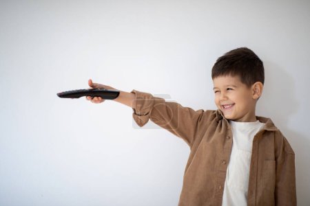 Foto de Cheerful small european boy with remote control plays, has fun, shoots on white wall background, free space. Entertainment with video on tv, game, fantasy, childhood at home, lifestyle at spare time - Imagen libre de derechos