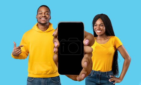 Foto de Mobile Ad. Smiling Black Man And Woman With Blank Smartphone In Hands Posing Isolated Over Blue Background, Happy African American Couple Recommending New App Or Website, Collage, Mockup - Imagen libre de derechos