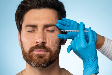 Foto de Hombre barbudo guapo de mediana edad que recibe inyecciones oculares en la clínica de belleza, fondo de estudio azul, primer plano. Hombre asistir esteticista, tener relleno de la cara - Imagen libre de derechos