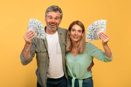 Téléchargez les photos : A lot of money, joy of victory. Happy middle aged caucasian couple holding many dollars and expressing emotions of success, isolated on yellow studio background - en image libre de droit