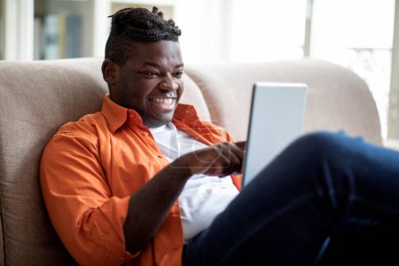 Téléchargez les photos : Happy cool plump young black guy with dreadlocks sitting on couch in living room, using digital tablet at home and smiling, chatting with friends, websurfing, using nice app, copy space - en image libre de droit