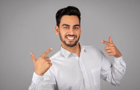 Téléchargez les photos : Smiling Arab Businessman Pointing Finger At Himelf And Looking At Camera, Handsome Self-Confident Middle Eastern Male Entrepreneur Standing Isolated Over Grey Studio Background, Copy Space - en image libre de droit