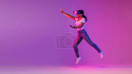Téléchargez les photos : Joyful Black Woman Wearing Vr Glasses Jumping On Air In Neon Light, Amazed African American Lady Experiencing Virtual Reality While Playing Video Game, Flying Over Purple Studio Background - en image libre de droit