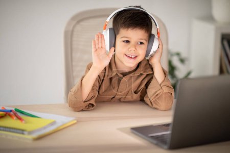 Téléchargez les photos : Smiling cute small european boy in headphones sitting at table, waving hand in laptop, has video call in room interior. Lesson at school, art, study, education at home remotely, childhood and device - en image libre de droit