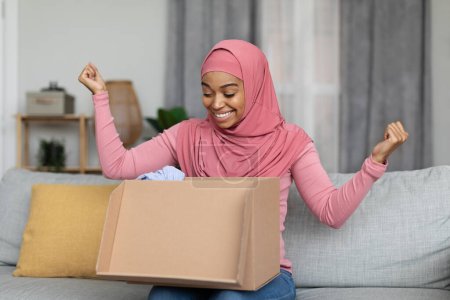 Photo for Online shopping and delivery banner. Overjoyed black muslim woman opening parcel and making YES gesture, satisfied with her ordered product - Royalty Free Image