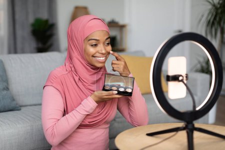 Téléchargez les photos : Heureuse femme musulmane noire maquilleuse assise devant un téléphone portable posé sur une lampe annulaire, en streaming depuis la maison tout en appliquant des cosmétiques, blogueuse beauté montrant une nouvelle palette à la caméra - en image libre de droit