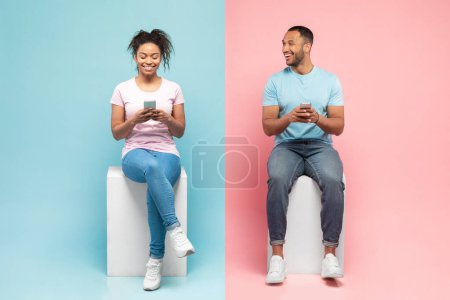 Téléchargez les photos : Excited african american couple sitting on halved pink and blue background, happy man looking aside at her girlfriend, using together smartphones - en image libre de droit