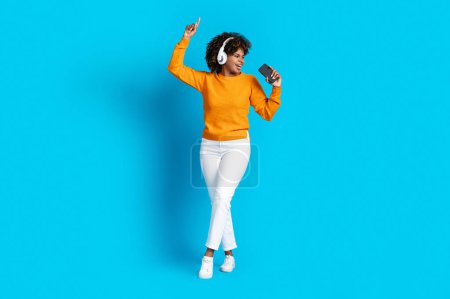 Foto de Happy stylish cute young black woman in casual outfit using modern wireless headphones and cell phone, singing and dancing, using phone as microphone, blue studio background, copy space, full length - Imagen libre de derechos