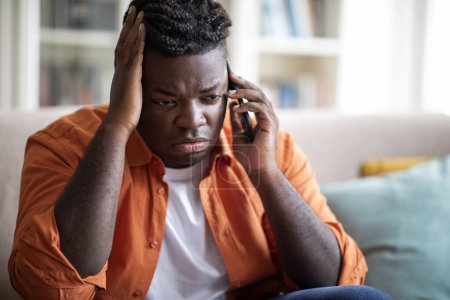 Photo for Closeup of stressed overweight black guy sitting on couch, having conversation on mobile phone, touching his head. Upset african american young man talking on smartphone, having bad news, copy space - Royalty Free Image