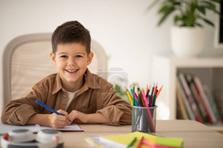 Téléchargez les photos : Smiling cute small european boy sitting at table, draws picture, look at camera in school, children garden room interior. Elementary education, art, study, learning at home and childhood lifestyle - en image libre de droit