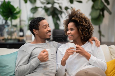 Téléchargez les photos : Beau jeune couple afro-américain en pyjama assis sur le canapé dans le salon confortable, tenant des tasses à café, avoir la conversation et rire, les amoureux de boire du café le matin à la maison - en image libre de droit