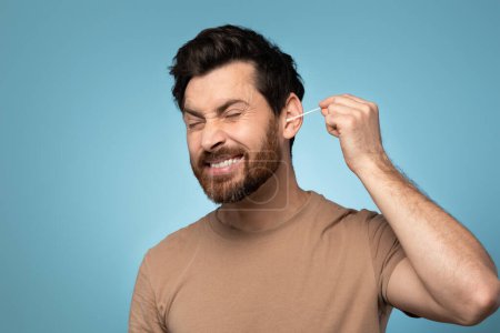 Téléchargez les photos : Daily hygiene routine. Funny bearded man cleaning ears with cotton swab while doing morning beauty routine, blue background. Clean ear and safe earwax removal - en image libre de droit