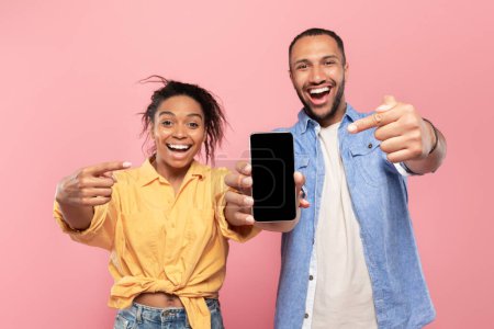 Foto de Utilice esta nueva aplicación. Emocionada pareja negra apuntando al teléfono inteligente con pantalla en blanco, recomendando aplicación móvil o sitio web de publicidad, maqueta para el diseño - Imagen libre de derechos