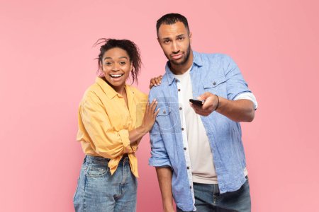 Photo for Black couple with remote control watching TV over pink studio background, young spouses with controller watching television program or show - Royalty Free Image
