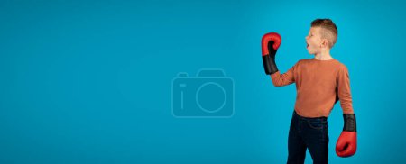 Photo for Emotional Boy Wearing Boxing Gloves Looking Aside At Copy Space, Sporty Preteen Male Child Raising Hand And Exclaiming With Excitement While Standing Isolated Over Blue Studio Background, Panorama - Royalty Free Image