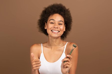 Téléchargez les photos : Cheerful millennial multiethnic woman show thumb up, jade roller isolated on brown background, studio. Recommendations for taking care of natural beauty, routine procedures and cosmetic spa treatments - en image libre de droit