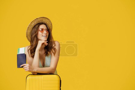 Téléchargez les photos : Happy stylish lady with passport and flight tickets leaning on suitcase and looking aside at free space, standing over yellow studio background - en image libre de droit