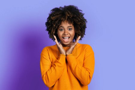 Téléchargez les photos : Surprised emotional cute attractive young black woman with bushy hair and teeth braces looking at camera and gesturing, purple studio background. Excitement, human emotions concept - en image libre de droit