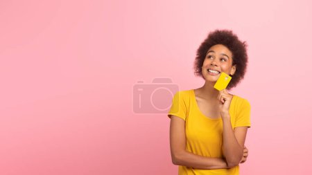 Foto de Feliz pensativo joven afroamericano rizado mujer shopaholic en camiseta amarilla con tarjeta de crédito pensar en compras, venta aislada sobre fondo rosa, estudio. Finanzas, dinero para compras, publicidad, oferta - Imagen libre de derechos