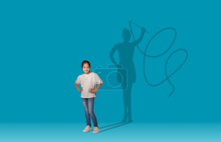 Téléchargez les photos : Ombre de gymnaste féminine avec bande sur cool rêveuse jolie asiatique écolière posant sur fond de mur studio bleu, espace de copie, collage. Concept d'enfance et de grands rêves - en image libre de droit