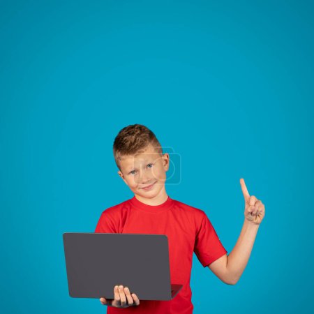 Téléchargez les photos : Smiling Preteen Boy Holding Laptop And Pointing Finger Up, Portrait Of Happy Male Child Having Idea While Study With Computer, Cheerful Lid Standing Isolated Over Blue Background, Copy Space - en image libre de droit
