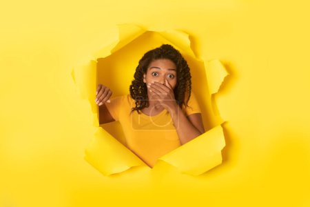 Téléchargez les photos : Incroyable nouvelle. Choquée femme afro-américaine fermant la bouche avec la main, regardant à travers un trou dans du papier jaune déchiré. Omg, grande offre, promotion cool, nouvelles étonnantes - en image libre de droit