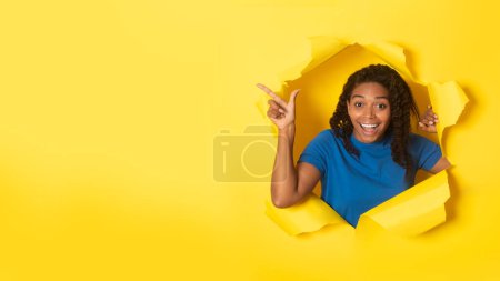 Téléchargez les photos : Overjoyed black woman pointing finger aside at free space and smiling at camera, posing in torn yellow paper background, panorama. Wow offer advertisement concept - en image libre de droit