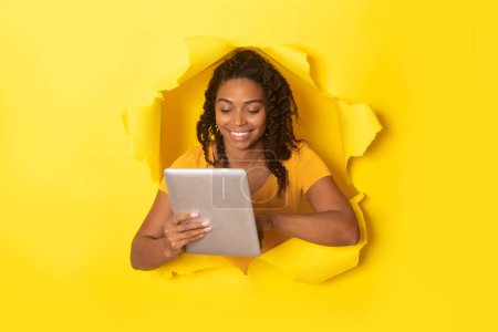 Foto de Positive black lady using digital tablet, posing in hole in torn paper on yellow background, studio shot, collage. Communication, great application concept - Imagen libre de derechos