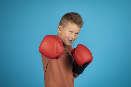 Téléchargez les photos : Sporty Male Kid Wearing Boxing Gloves And Making Punch At Camera, Cute Preteen Boy Standing In Fighter Posture, Ready To Fight, Posing Isolated Over Blue Studio Background, Copy Space - en image libre de droit