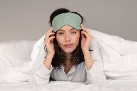 Photo for Calm pretty caucasian millennial female in sleep mask lies on bed under white blanket, enjoys weekend, vacation and free time in bedroom interior. Health care, sweet dreams and good morning at home - Royalty Free Image