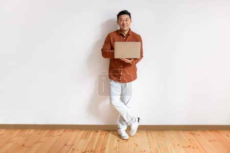 Photo for Full length portrait of positive middle aged asian man using laptop computer for online work, education or communication, standing against white studio wall, copy space - Royalty Free Image